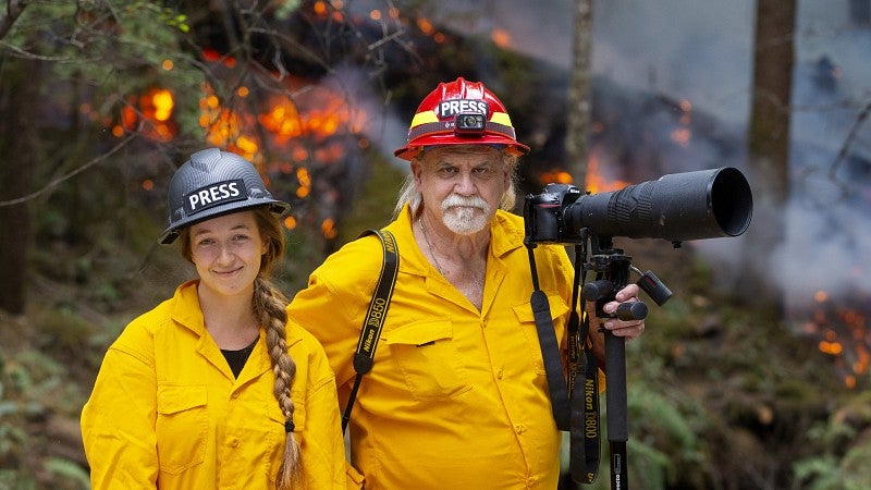 a student and Dan Morrison wearing hard hats labeled Press and yellow jumpsuits pose with a camera in front of a wildfire
