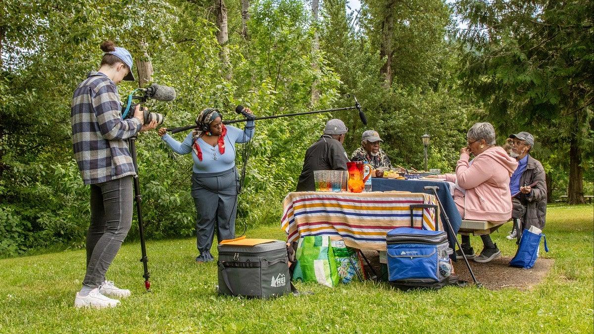 two people using video and sound recording equipment film a group of Black Portlanders having a picnic in a park