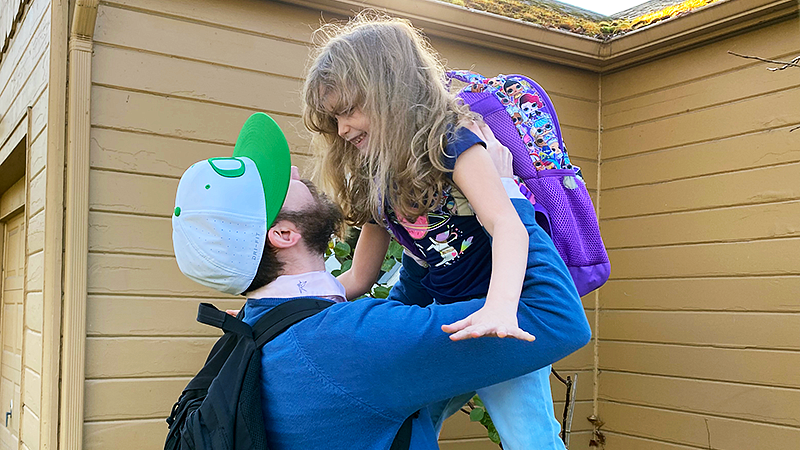 Johnny Media wears a white and green UO cap and holds his daughter in the air