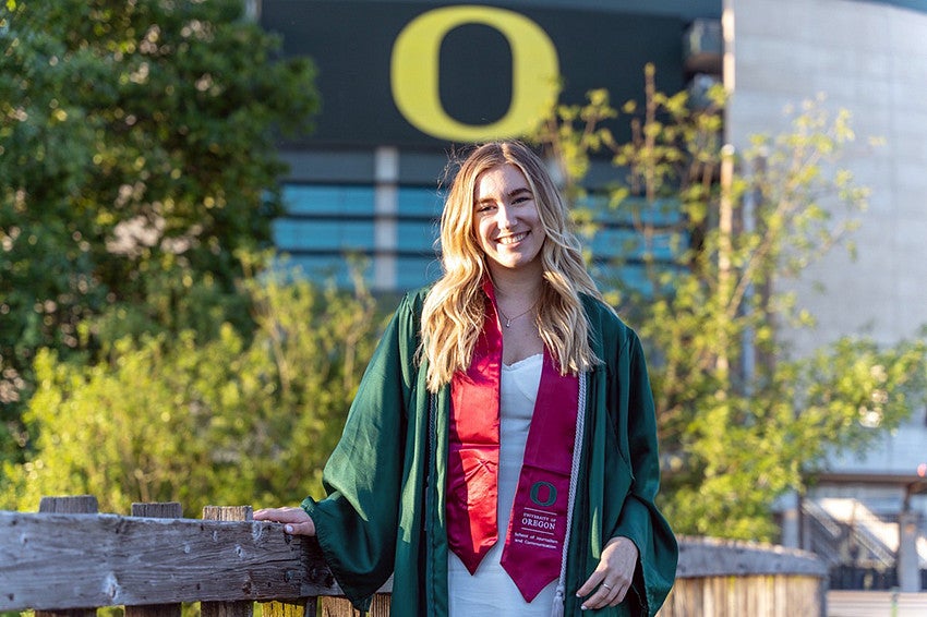 Haley Landis wears a graduation gown and burgundy SOJC stole with the yellow O at Autzen stadium in the background