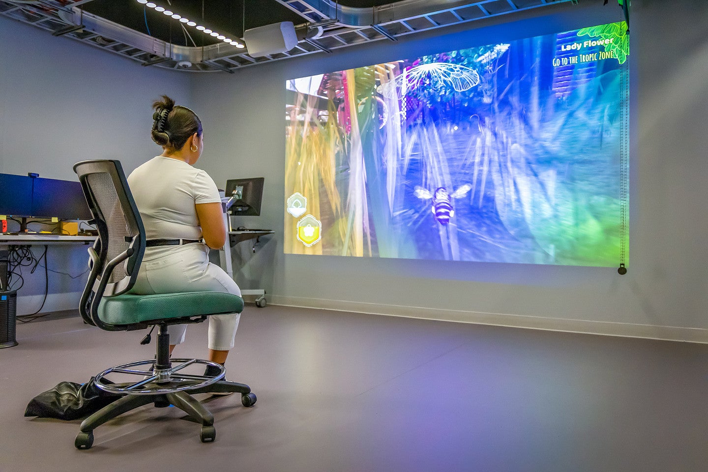 a student sits on a rolling chair facing a large screen showing a vibrant video game
