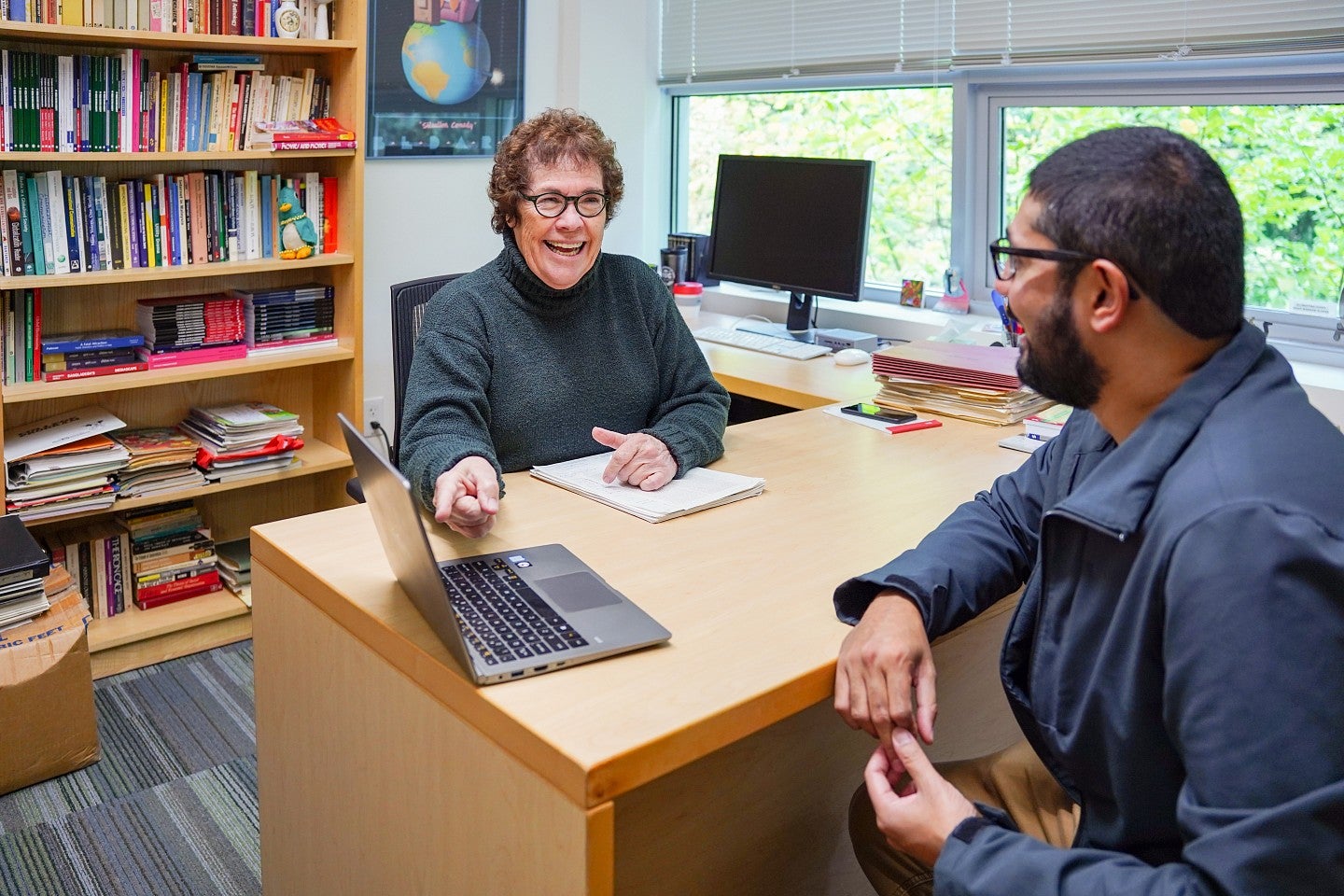 janet wasko sits at her desk and gestures to a laptop while smiling at a student