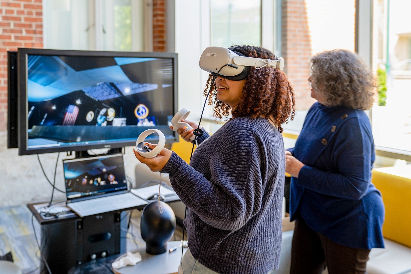 a student uses a VR headset in front of a screen showing the metaverse while Donna Davis looks on