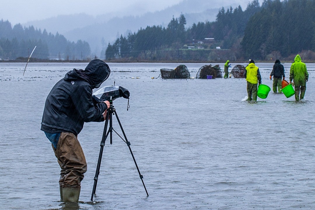 a student wearing rain gear films fishermen while standing in a shallow body of water