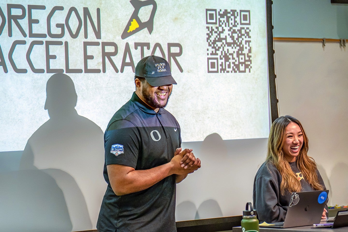 Justin Johnson and Christina Nguyen present in front of a projector screen that says Oregon Accelerator