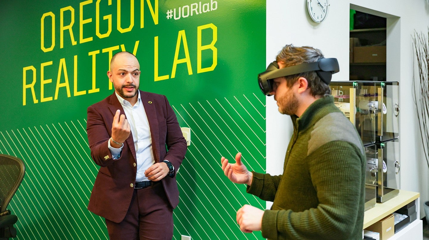 Danny Pimentel gestures to a student wearing a VR headset in front of a green wall that says Oregon Reality Lab