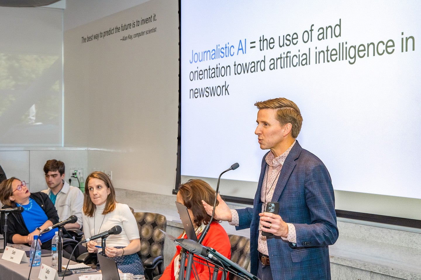 Seth Lewis presents at a microphone from a panel of experts. A screen behind him says "Journalistic AI = the use of and orientation toward artificial intelligence in newswork."