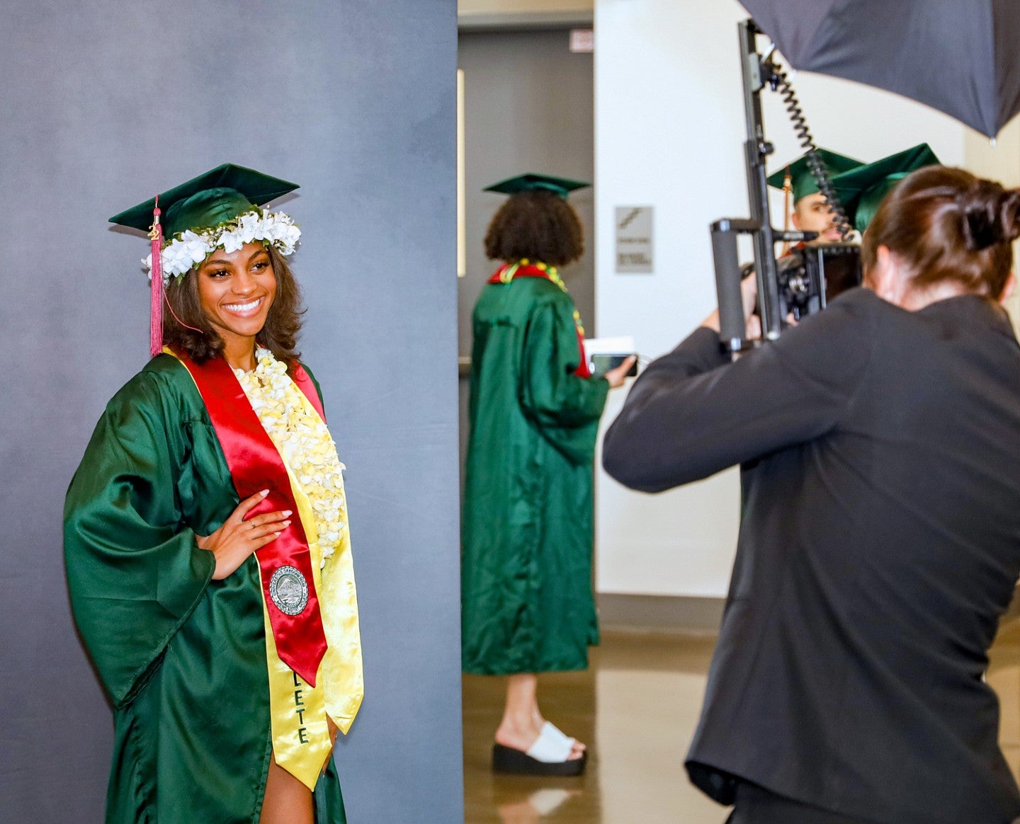 a photographer takes a photo of a student wearing commencement regalia and posing in front of a backdrop