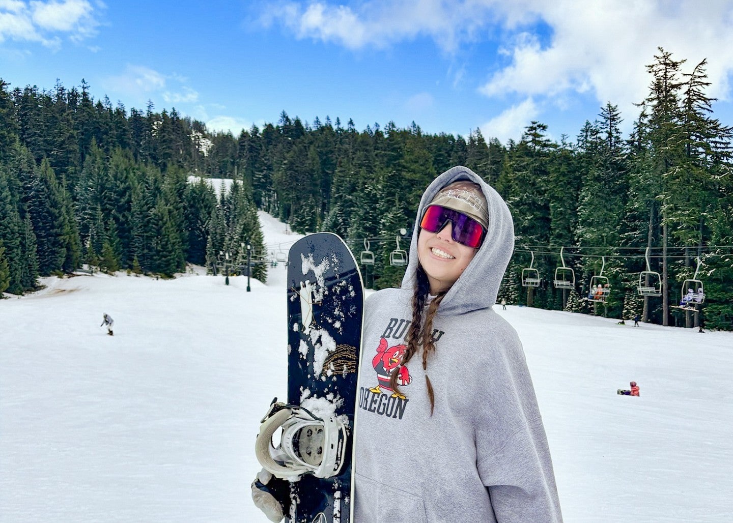 Sydney Seymour wears ski goggles and holds a snowboard while standing in front of a ski lift rising amid evergreen trees