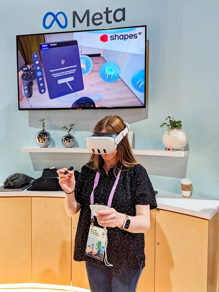 Kathleen Darby wears a VR headset in front of a wall with the Meta logo and a large screen showing an immersive environment