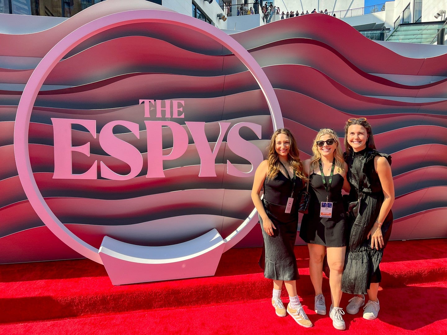 Three sports PR professionals pose on a red carpet at the Espy awards, with a large 3D logo on an undulating red backdrop behind them