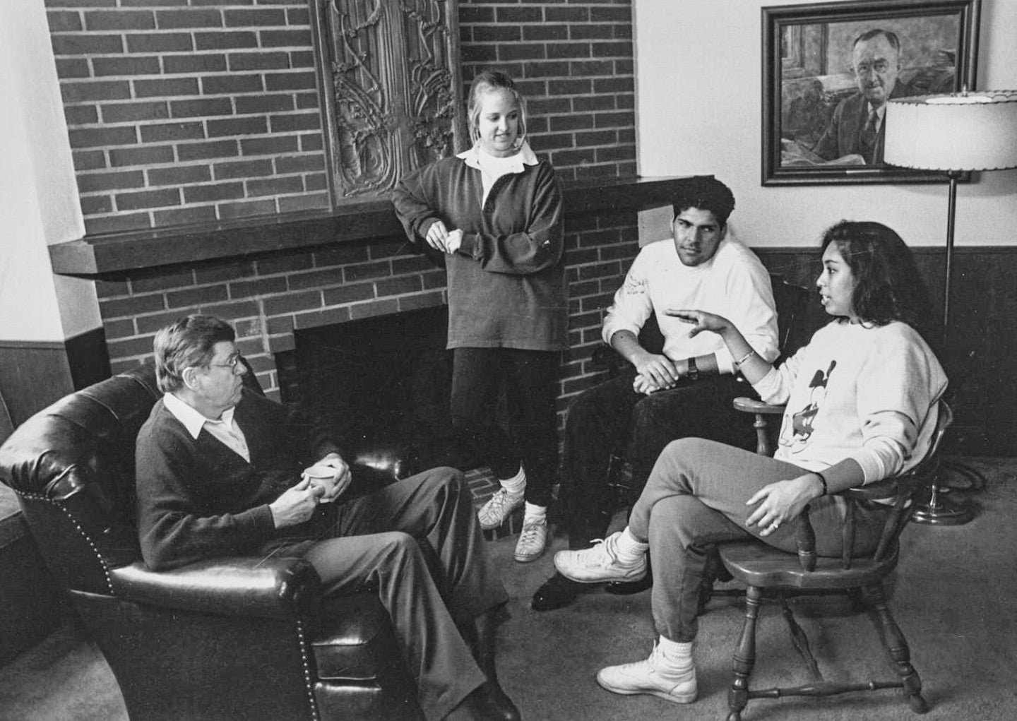 black and white historic photo of students and professor having a conversation in front of the Allen Room fireplace