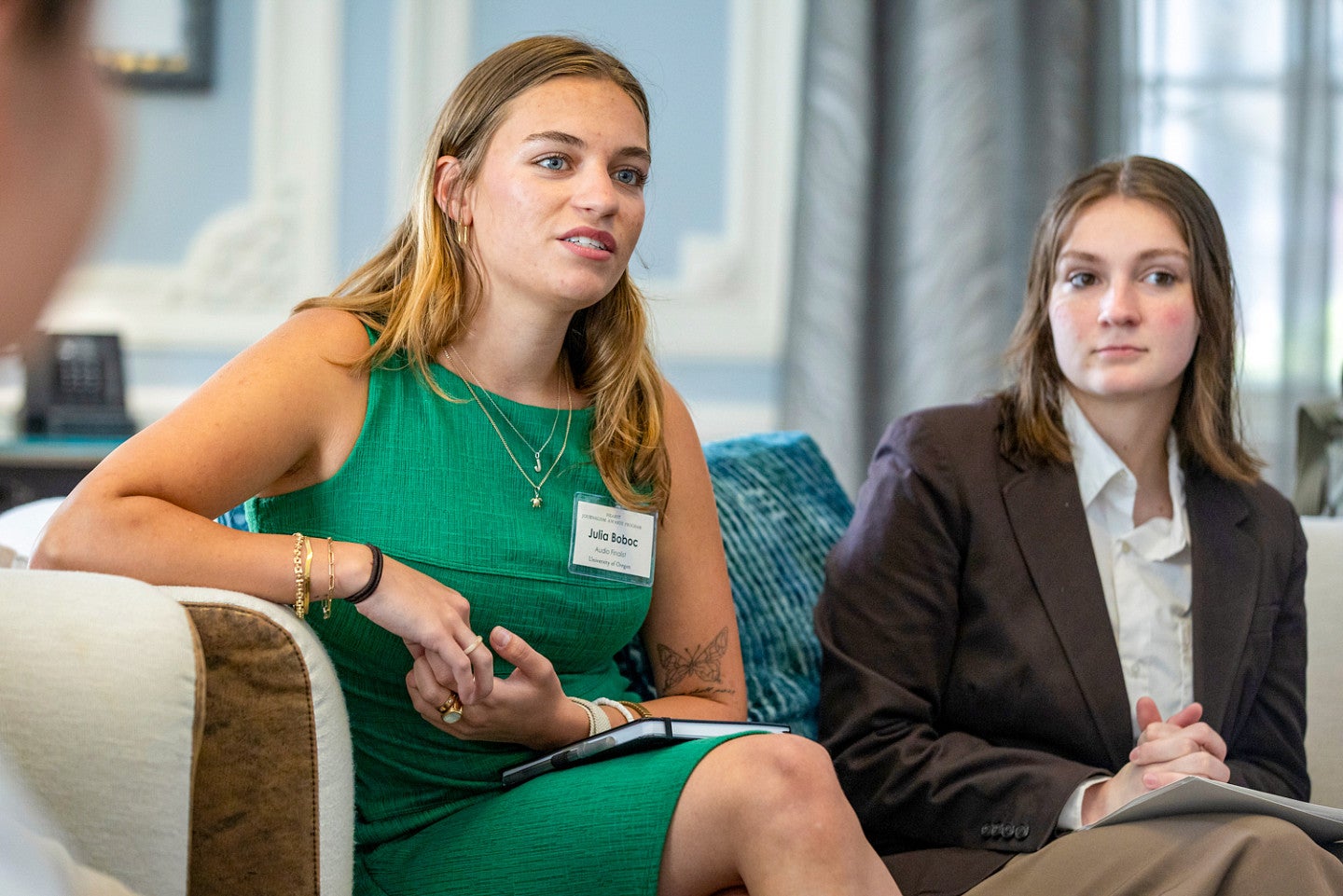 Julia Boboc is wearing a green dress and sitting on a couch while talking to someone out of frame. Another unidentified student sits next to her.