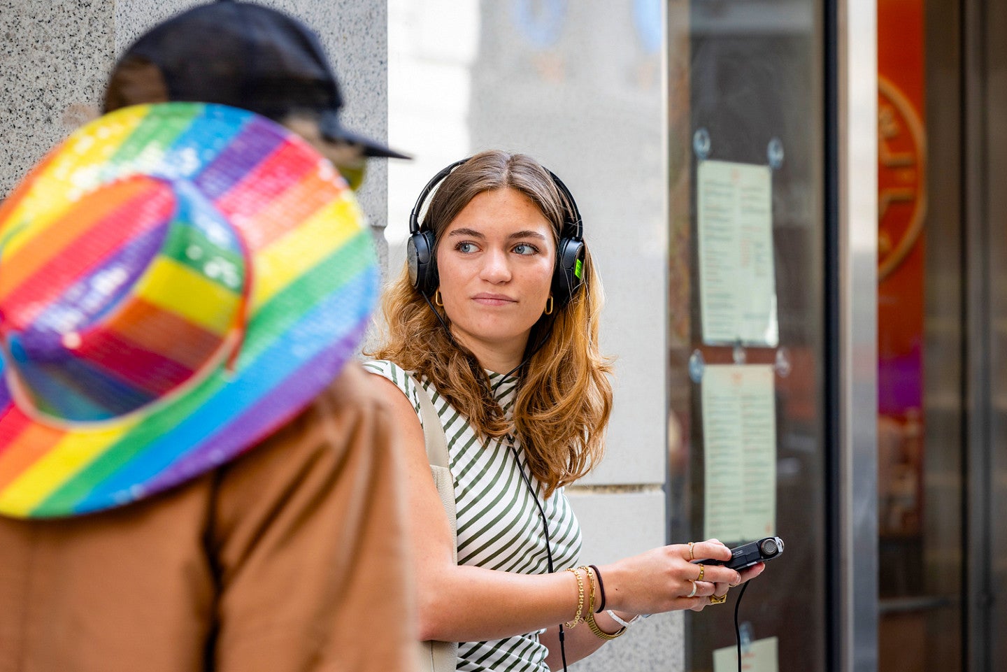 Julia Boboc wears headphones and holds a recorder while standing on a sidewalk. She is in focus in the middle of the frame; in the foreground, a person with a rainbow striped hat is about to walk past Julia.