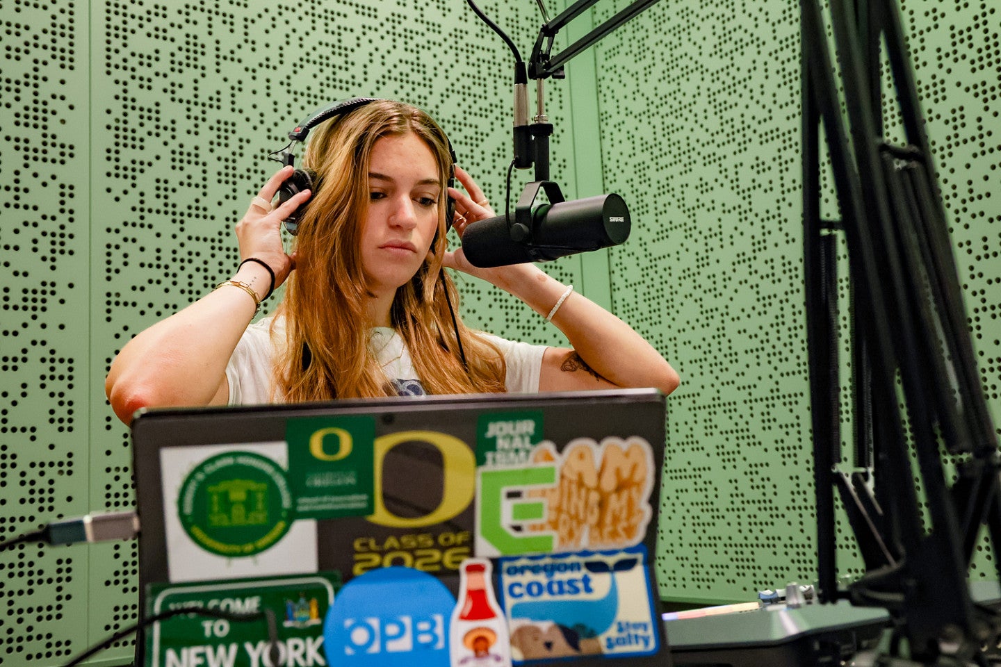 Julia Boboc adjusts her headphones while sitting in front of a microphone and her computer in a podcast studio with walls lit in green