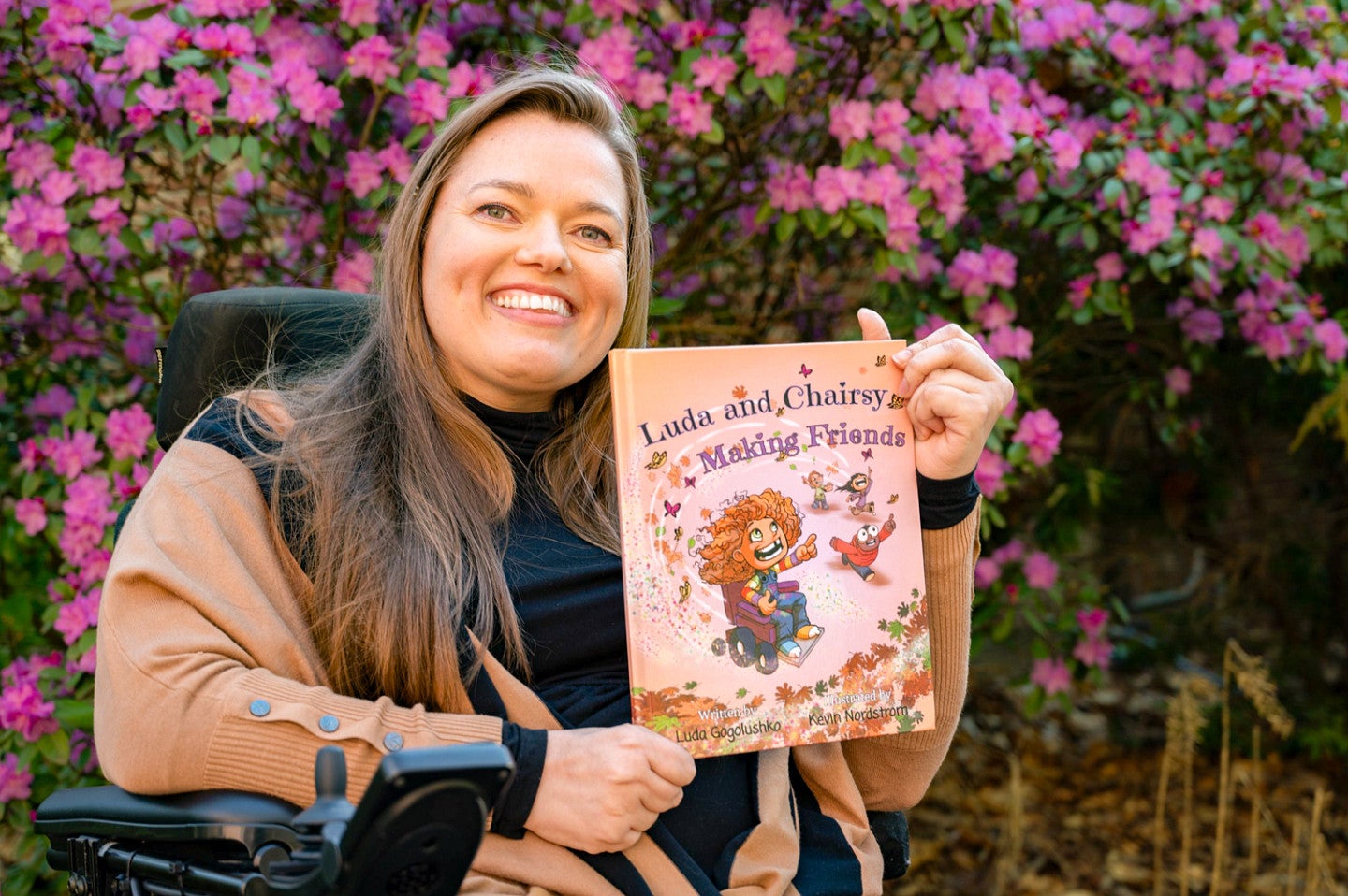 Luda Gogolushko holds up her children's book, titled "Luda and Chairsy Making Friends;" Luda is sitting in her wheelchair in front of a wall of fuschia flowers 
