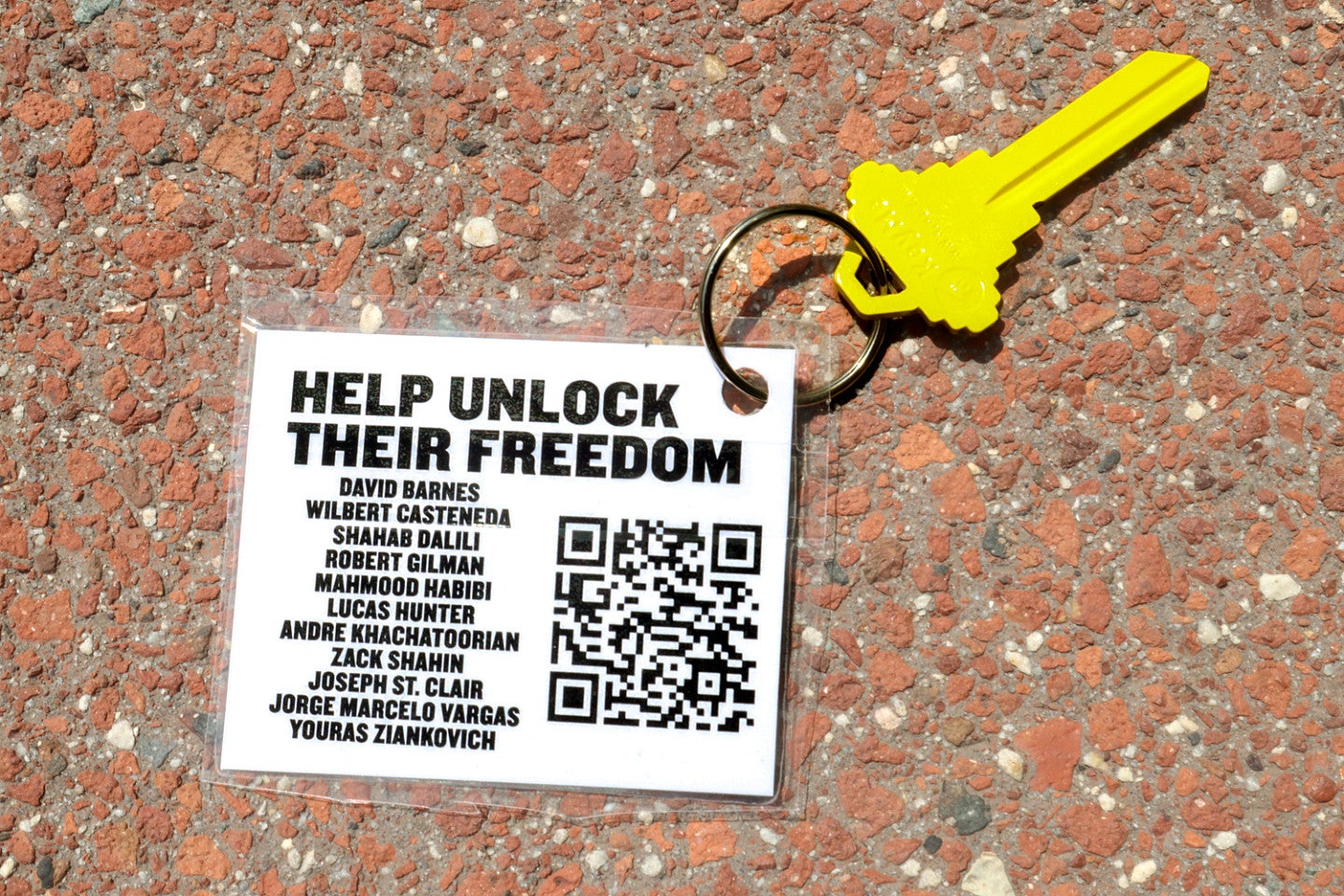A bright yellow key attached to a white tag that says "Help Unlock Their Freedom" with a list of names and a QR code 