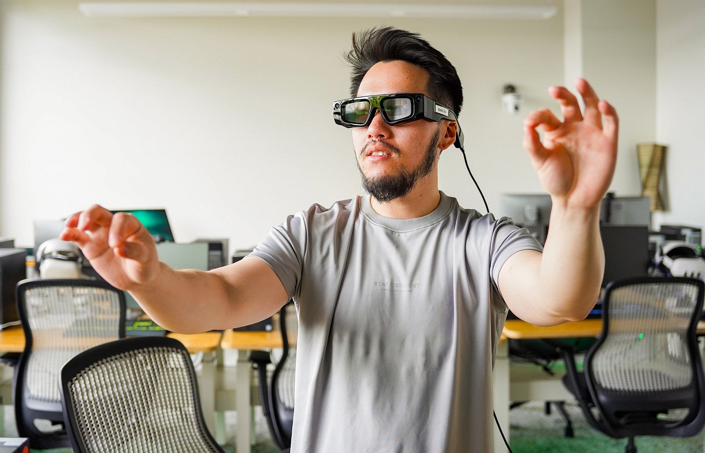 A student with dark hair and a goatee wears augmented reality glasses and motions with his hands while in a computer lab