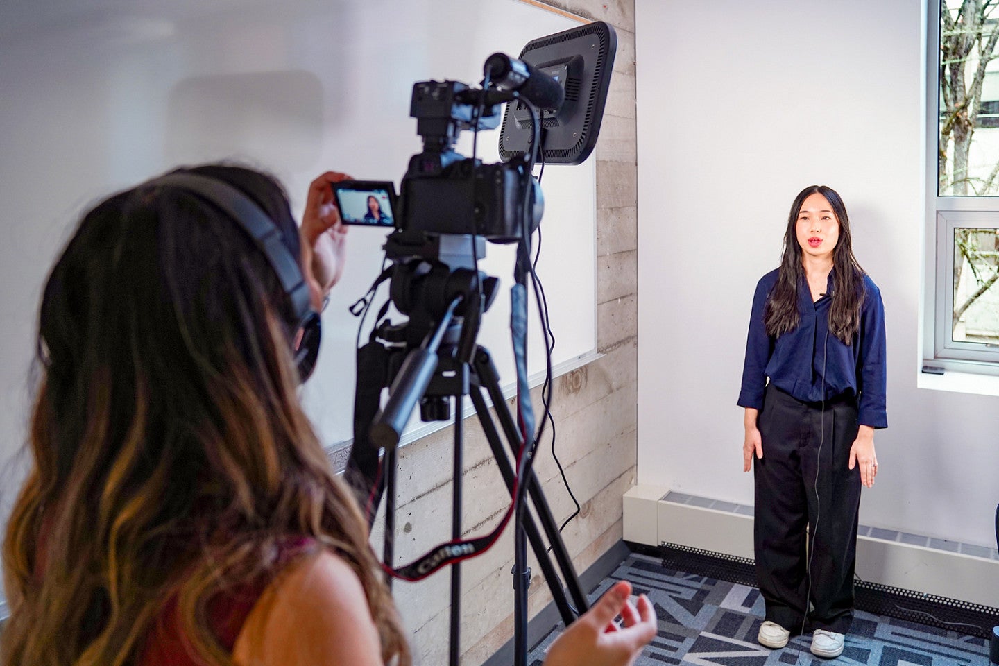 Cing Dim stands behind a video camera filming Mary Pham in a classroom setting
