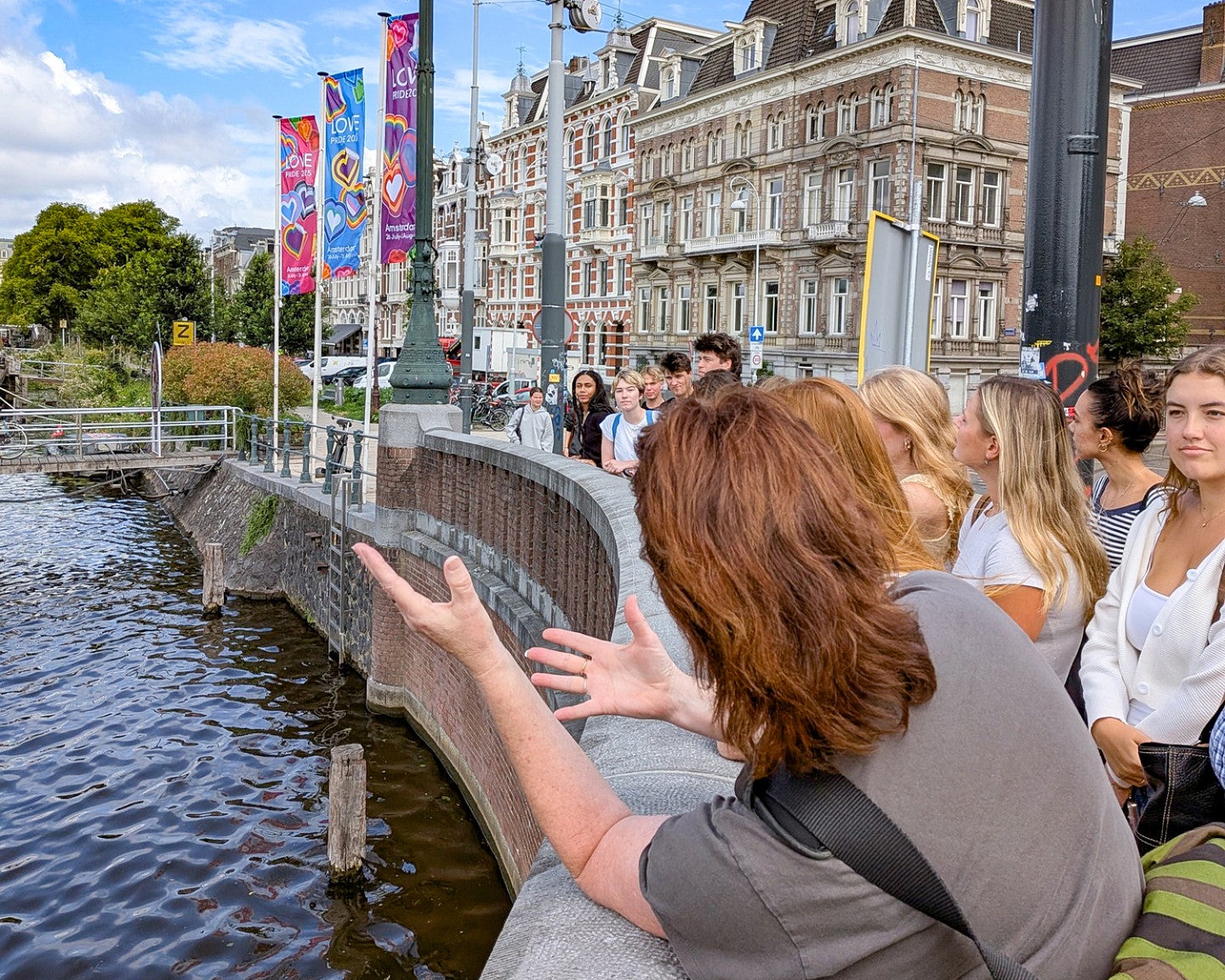 a group of students look out on to a river from a stone bridge with a person gesturing in the foreground, seen from behind