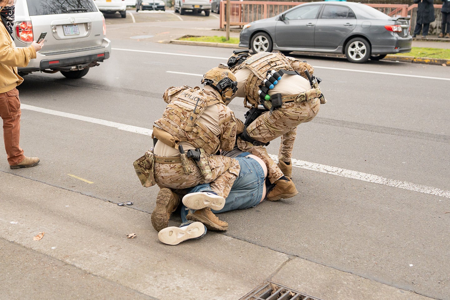 two uniformed ICE officers pin a civilian protestor down with their knees on the street with cars passing in the background