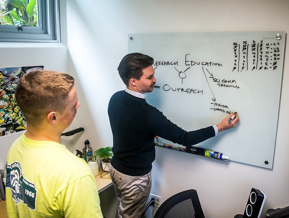 Two people work together in an office space. One person writes on a white board while the other person observes. 