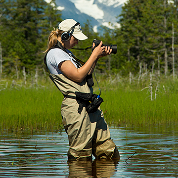 a student wearing headphones and holding a camera stands in a mountain river