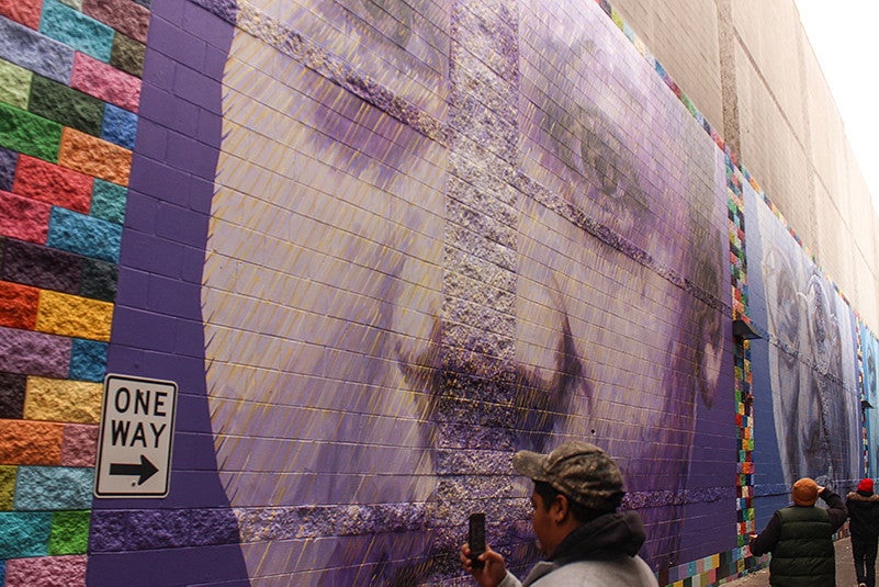 A person wearing a camouflage hat takes a photo of a purple mural.