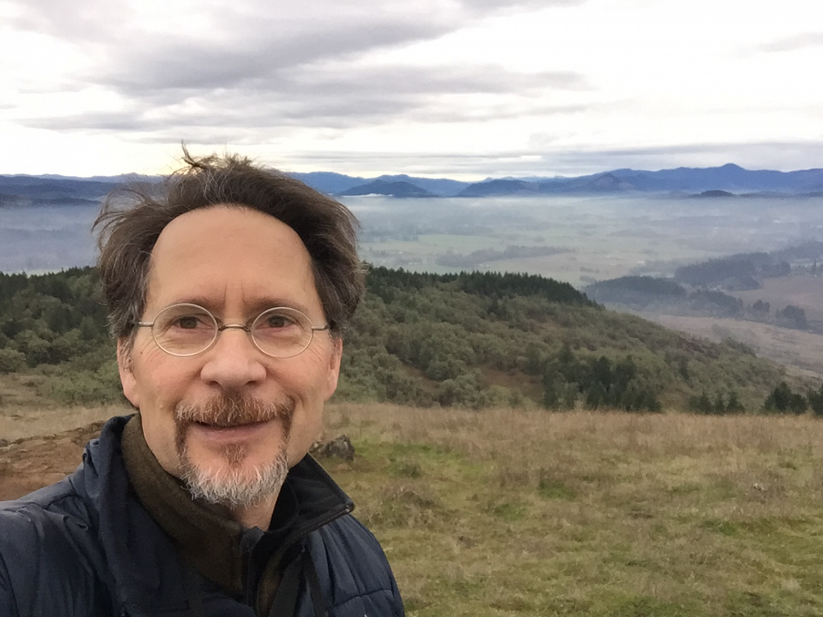 Chris Frisella takes a selfie in front of a cascading mountain view.