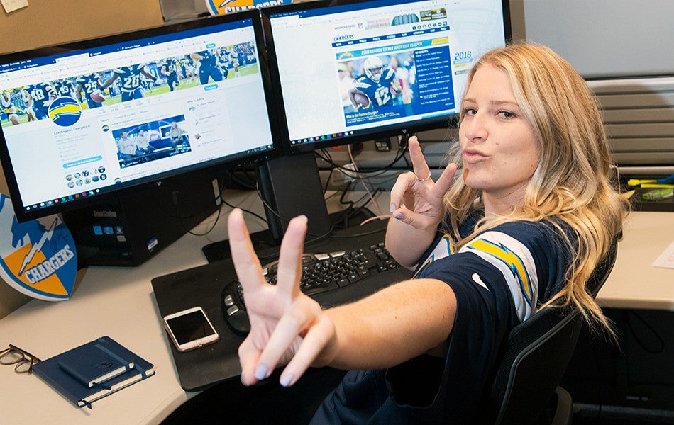 A female student sitting behind a bank of computer monitors giving the peace sign to the camera