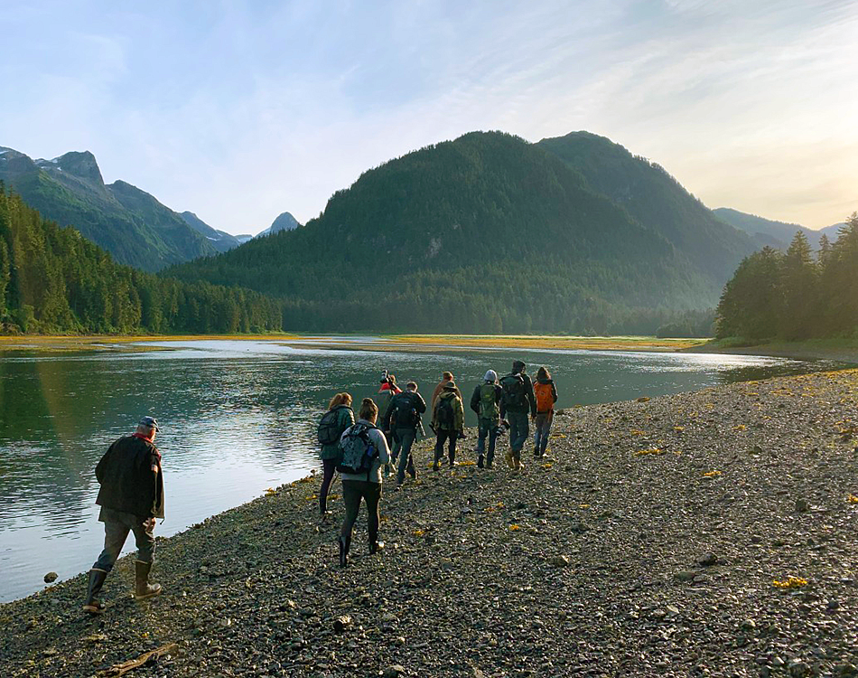 A group of people with their backs to the camera walk along a river with a mountain scene at sunrise behind them