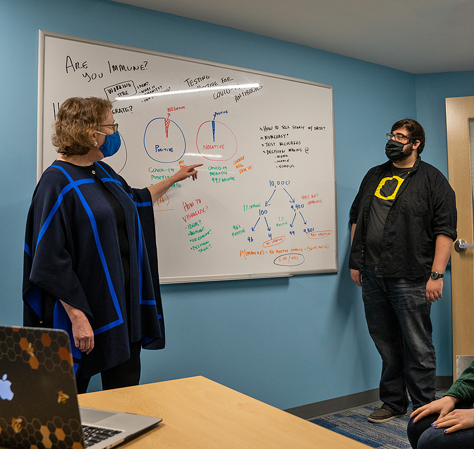 SCR director Ellen Peters gestures to a white board covered in notes about COVID-19 research while a student looks on