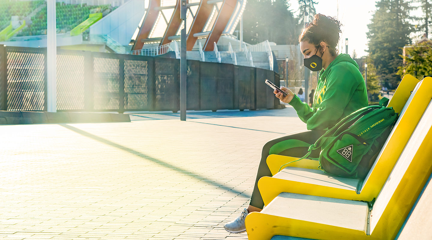 Journalism junior Peyton Brooks, dressed in green, sits on a yellow bench outside the newly rebuilt Hayward Field.  