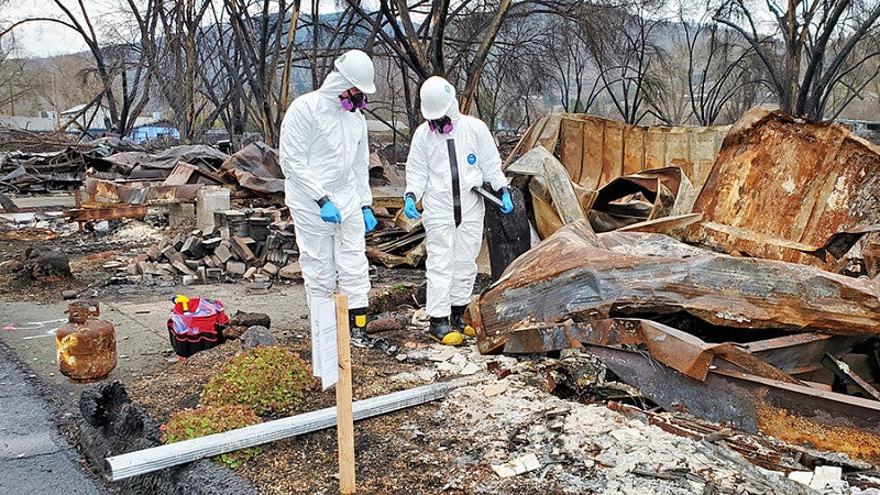 two people wearing white hazmat suits inspect a property destroyed by fire