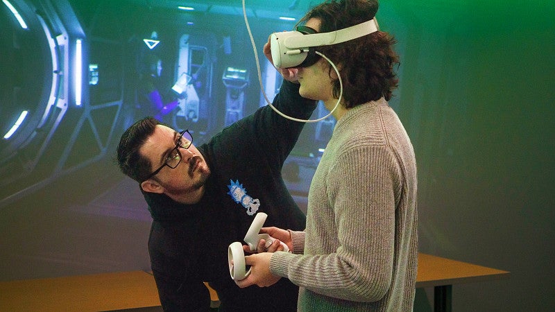 two people work with VR equipment in front of a screen displaying the VR content