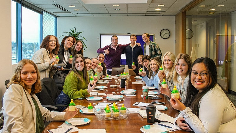 several people are gathered around a conference room table holding cupcakes with green icing and small yellow ducks on top