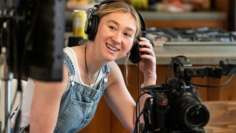 a student using camera equipment adjusts headphones while smiling at a monitor 