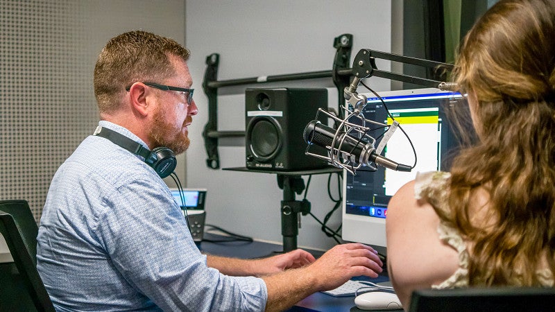 Charlie Deitz works at a computer in the SOJC podcast studio while talking to a student