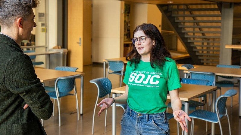 A student wearing a green SOJC tee shirt speaks with someone in the atrium of Allen Hall