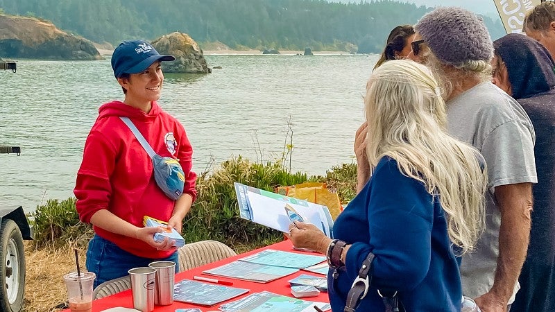 Sof Fox, a UO science communication minor, stands at a table laid out with informational materials with a river and pine forest in the background