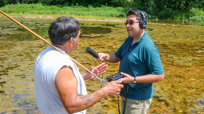 Brian Bull records an audio interview with another person while standing in a shallow murky pond