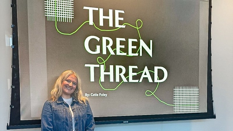 Catie Foley stands in front of a screen displaying the title of her Advertising and Brand Responsibility master's capstone project called "The Green Thread"