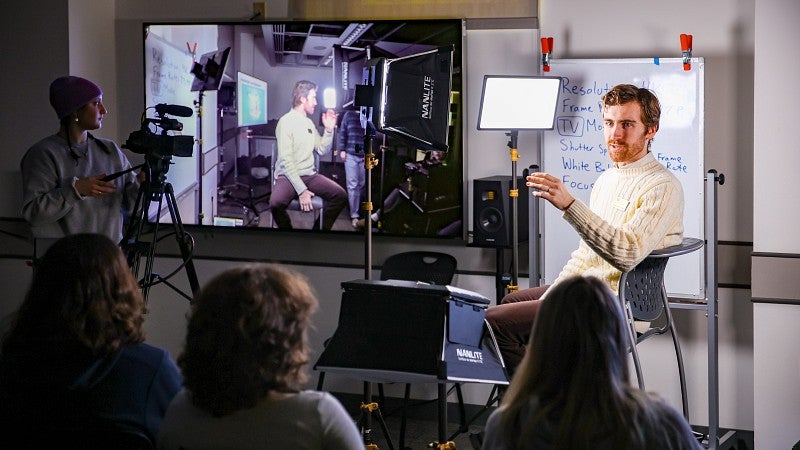 an instructor sits in front of students while a screen showing video of him is next to him