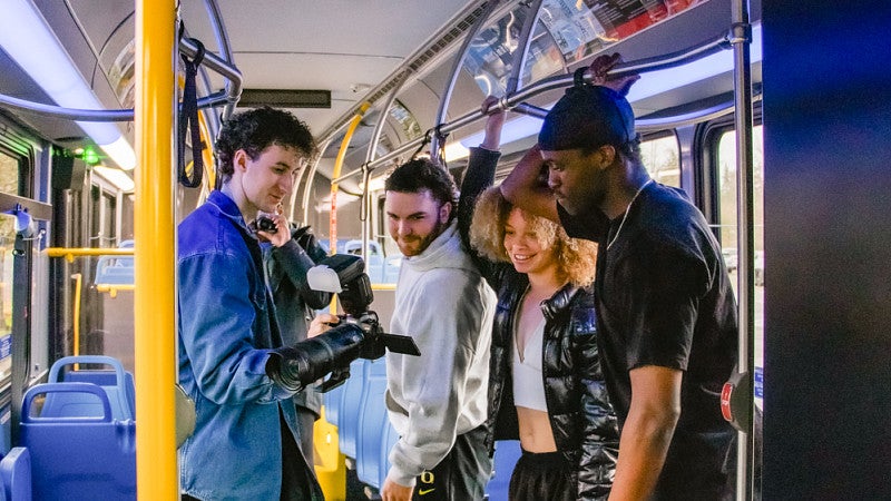 a student photographer shows three student athletes the back of his camera during a photo shoot on board a bus