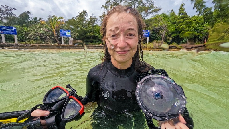Bea Mira grins at the camera while wearing a wetsuit, holding camera gear and standing in the ocean