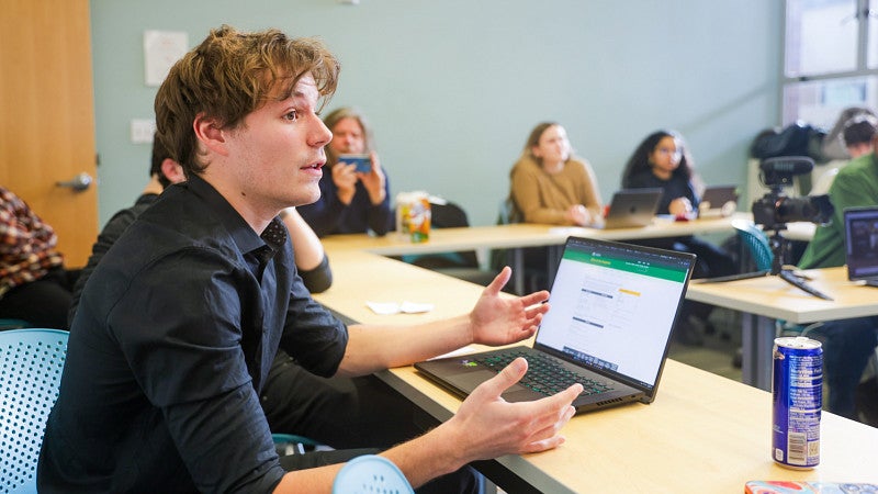 a student in the foreground speaks to someone off camera; other students in the background sit around the table