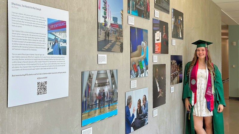 Ally Thomas, wearing her graduation cap and gown, stands next to her photo exhibit in Allen hall