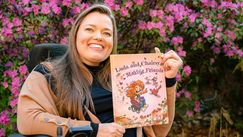 Luda Gogolushko holds up her children's book, titled "Luda and Chairsy Making Friends;" Luda is sitting in her wheelchair in front of a wall of fuschia flowers 
