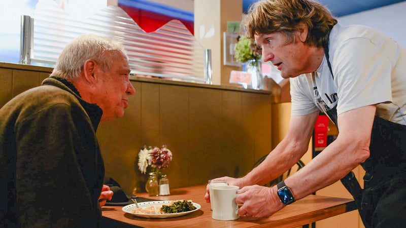 a person wearing an apron leans down to speak to another person sitting in a restaurant booth
