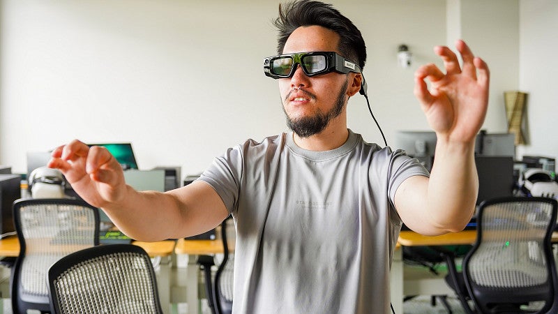 A student with dark hair and a goatee wears augmented reality glasses and motions with his hands while in a computer lab